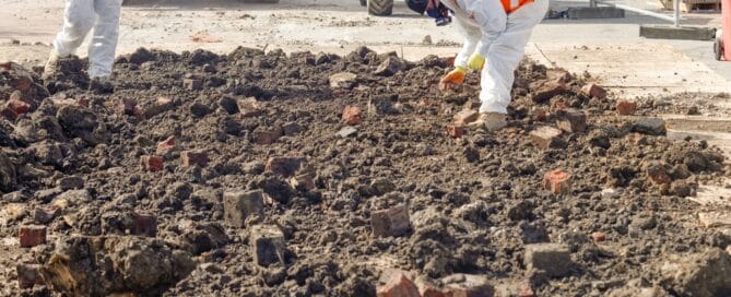 Two Workers In Protective Gear Remove Asbestos From Contaminated Ground